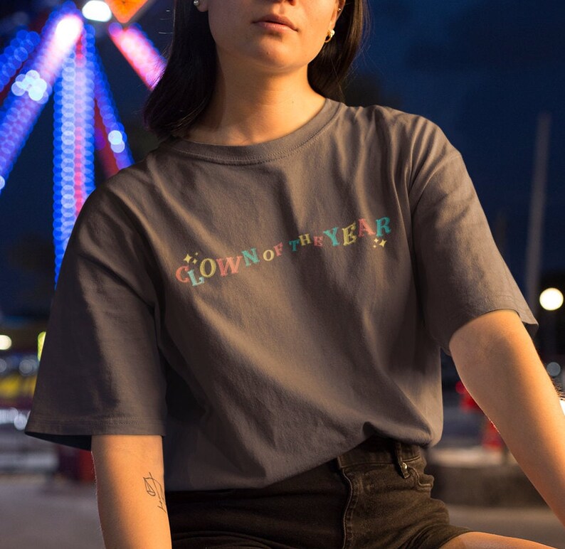 May include: A dark gray t-shirt with the text "CLOWN OF THE YEAR" in rainbow colors. The text is in a curved arc with stars on either side.