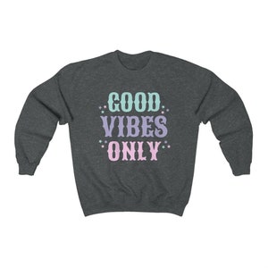 May include: A dark heather grey sweatshirt with the text "GOOD VIBES ONLY" in pastel pink, blue, and purple lettering with white stars around the text.