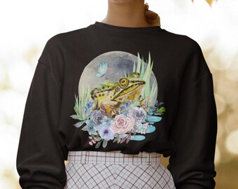 May include: Black sweatshirt featuring a watercolor frog design. The frog is green and yellow, surrounded by blue, pink, and purple flowers, with a butterfly and a moon in the background. The design is centered on the front of the sweatshirt.