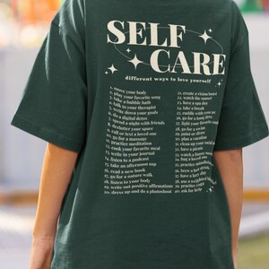 May include: A green t-shirt with the text "SELF CARE" in a white circle with stars. The text "different ways to love yourself" is below the circle. A list of 40 self-care tips is printed on the back of the shirt.
