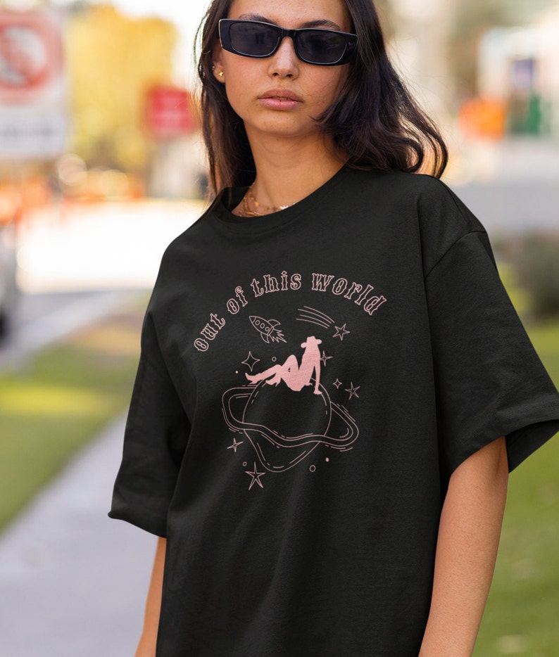 May include: Black t-shirt with a pink graphic of a woman sitting on a planet with the text "out of this world" above her.