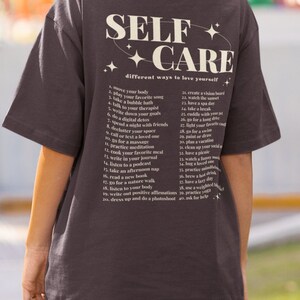 May include: A dark gray t-shirt with the text "SELF + CARE" in white and a list of self-care tips in a smaller font. The text reads "different ways to love yourself".