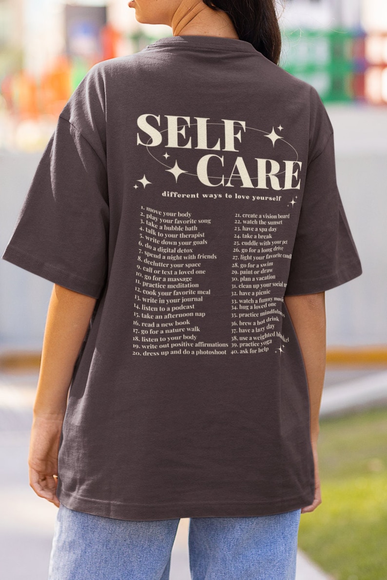 May include: A dark gray t-shirt with the text "SELF + CARE" in white and a list of self-care tips in a smaller font. The text reads "different ways to love yourself".