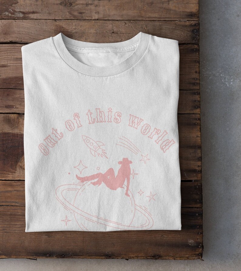 May include: White t-shirt featuring a pink graphic design of a woman sitting on a planet with a rocket ship and stars. The text "out of this world" is written in a pink cursive font.