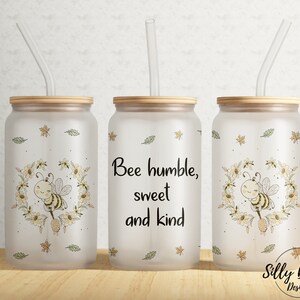 Può includere: Tre bicchieri in vetro smerigliato con coperchi in bambù e cannucce. Ogni bicchiere presenta un design diverso: un'ape con una ghirlanda di fiori, il testo "Bee humble, sweet and kind", e un'ape con una ghirlanda di fiori.