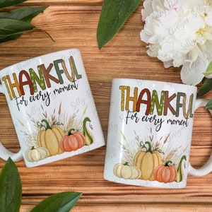 May include: Two white ceramic mugs with a fall theme. The mugs feature the text "Thankful for every moment" with a colorful, patterned font. The mugs are decorated with a fall scene of pumpkins, gourds, and wheat stalks.