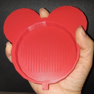 May include: Red plastic mold with a circular indentation and two ear-shaped protrusions. The mold has a textured surface.