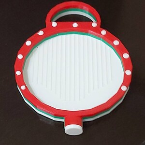 Op de afbeelding: Een rode, witte en groene kerstbal met een wit handvat en witte stippen rond de rand. De bal is gemaakt van plastic en heeft een getextureerd oppervlak.
