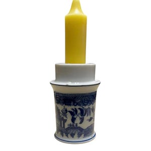 May include: A white ceramic candle holder featuring a blue willow pattern with birds and architectural motifs. The cylindrical base has a flared rim and holds a single, smooth, yellow pillar candle that extends vertically from the center.