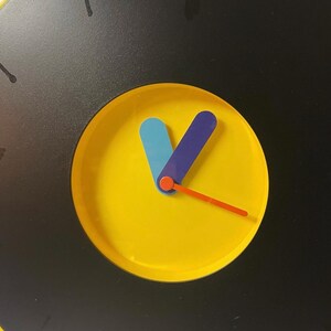 May include: A round wall clock with a black face and a yellow center. The clock hands are blue, purple, and orange. The clock has black markings for the hours.