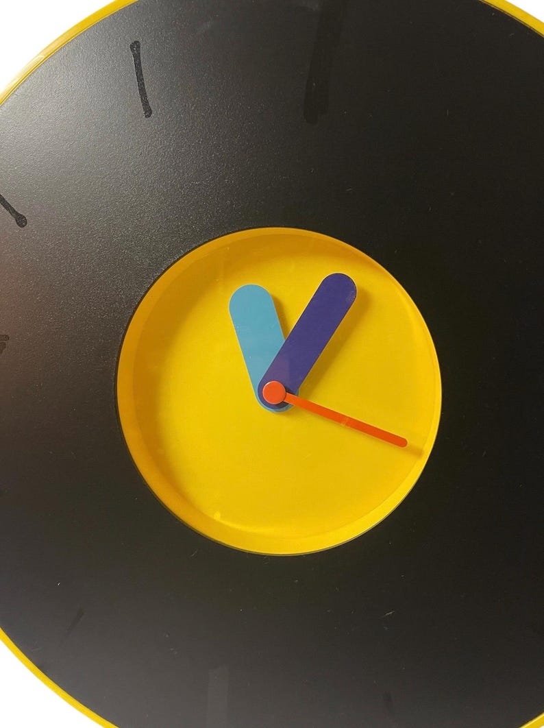 May include: A round wall clock with a black face and a yellow center. The clock hands are blue, purple, and orange. The clock has black markings for the hours.
