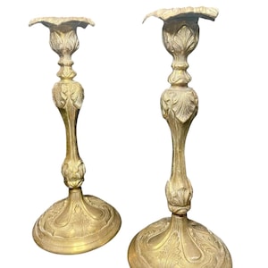 May include: Two antique gold-colored candle holders. Each holder has a round base with an ornate leaf design, a slender stem, and a flared top. The candle holders are made of metal and have a vintage aesthetic.