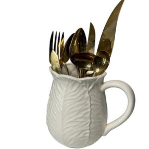 May include: White ceramic pitcher shaped like a cabbage with a handle. The pitcher is filled with gold-toned silverware, including a fork, knife, and spoons.