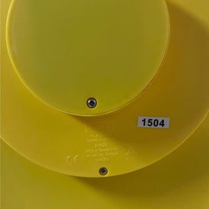 May include: Close-up of a bright yellow plastic object with a circular design. The object has a sticker with the number "1504". The object has a screw in the center and text that reads "PLADDA Made in China 21603 IKEA of Sweden AB SE-343 81 &Auml;lmhult PA66".