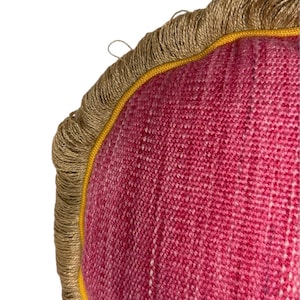 Puede incluir: Una almohada redonda de color rosa con ribete dorado y flecos marrones.