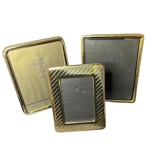 May include: Three gold-colored picture frames. The largest frame is 8x10 inches and has the text "SOLID BRASS Photo Frame hand polished 24 karat gold" on the front. The other two frames are smaller and have a textured design.