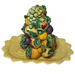May include: A ceramic fruit bowl with a yellow base and a stack of fruit on top. The fruit includes lemons, oranges, grapes, and plums. The fruit is painted in realistic colors.