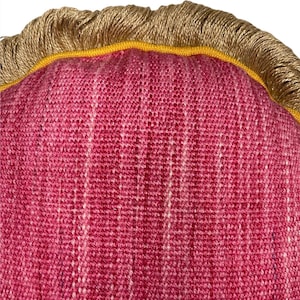 Puede incluir: Primer plano de una almohada tejida de color rosa y blanco con ribete de flecos dorado y amarillo.