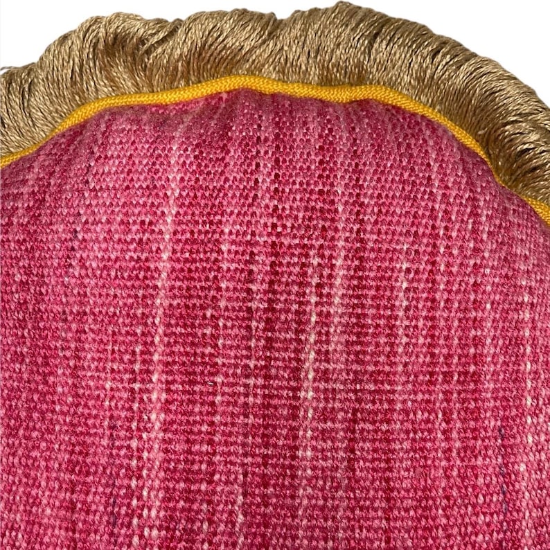 Puede incluir: Primer plano de una almohada tejida de color rosa y blanco con ribete de flecos dorado y amarillo.