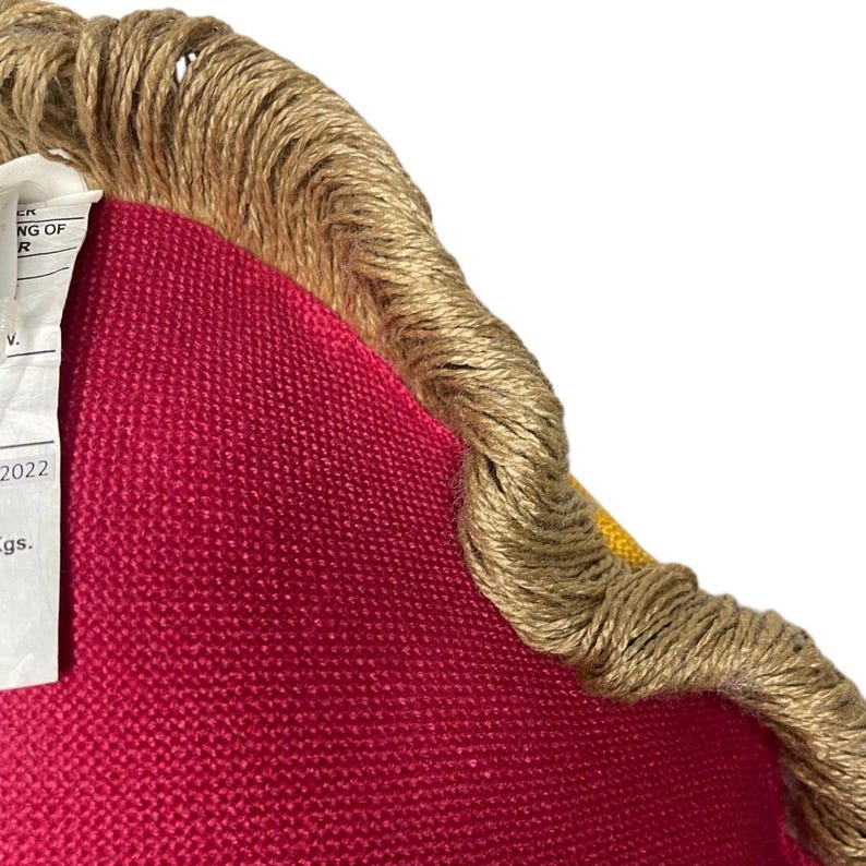 Puede incluir: Una almohada roja y amarilla con un ribete de cuerda marr&oacute;n. La almohada tiene una etiqueta adjunta con el texto "2022".