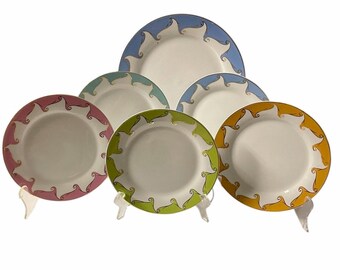 May include: A collection of six white ceramic plates with colorful rims. The rims feature a repeating swirl design in shades of pink, blue, green, and yellow. The plates are displayed on clear stands, showcasing their decorative edges.