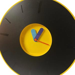 May include: A round wall clock with a black face and yellow trim. The clock has a yellow center with blue and purple hands and an orange second hand. The clock has a yellow loop at the top for hanging.
