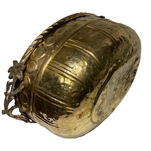 May include: A large, oval-shaped brass planter with a hammered finish. The planter has a decorative rim with a rope-like design and a floral motif on the handle.