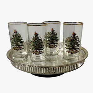 May include: A set of four clear glass tumblers with gold rims, each featuring a colorful illustration of a decorated Christmas tree. The glasses are arranged on a round, silver-toned metal serving tray with a decorative pierced gallery edge.