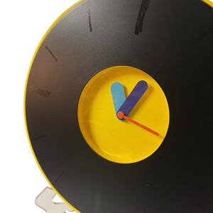 May include: A round, black and yellow clock with a vinyl record design. The clock face is yellow with blue and purple hands and an orange second hand. A yellow loop is at the top for hanging. The clock has a clear plastic stand.