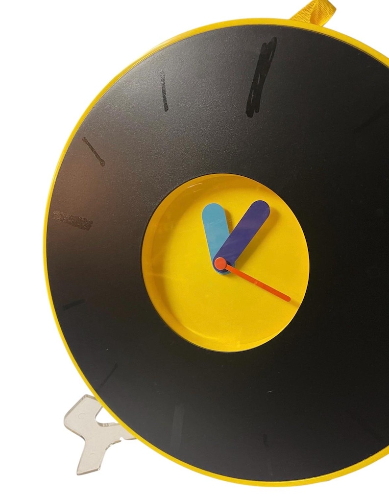 May include: A round, black and yellow clock with a vinyl record design. The clock face is yellow with blue and purple hands and an orange second hand. A yellow loop is at the top for hanging. The clock has a clear plastic stand.