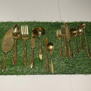 May include: A set of gold-toned silverware, including forks, spoons, and knives, laid out on a green artificial grass surface.