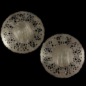 May include: Two silver-colored, circular coasters with intricate, openwork floral and leaf patterns around the edges. The center of each coaster has a brushed metal finish. The coasters are set against a black background.