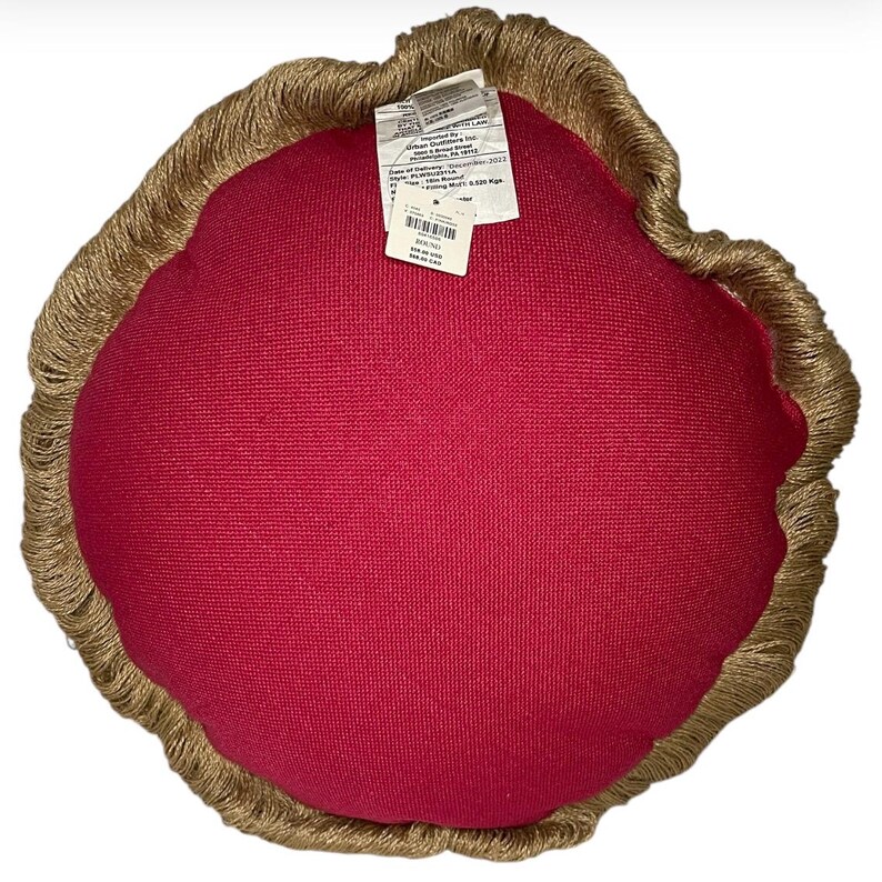 Puede incluir: Una almohada redonda de color rojo con un borde de flecos marrones. La almohada tiene una etiqueta adjunta con el texto "ROUND" y otra informaci&oacute;n.