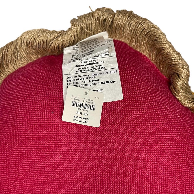 Puede incluir: Una almohada redonda de color rojo con un ribete de yute marr&oacute;n. La almohada tiene una etiqueta que dice "ROUND $58.00 USD $68.00 CAD".