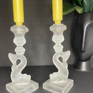May include: A pair of frosted glass candle holders shaped like fish. Each holder has a square base and a detailed design of a fish with its tail curled up. The holders are empty and ready to be used with candles.
