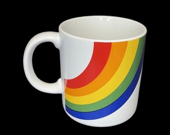 May include: A white ceramic mug featuring a curved rainbow graphic on the side. The design consists of four distinct bands of color in red, orange, yellow, green, and blue, arranged in a semi-circular arc pattern against the plain white surface.