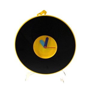 May include: A unique wall clock designed to resemble a vinyl record. The clock features a black disc with a yellow center and a yellow frame. The clock hands are blue and orange. The clock is supported by a clear stand.