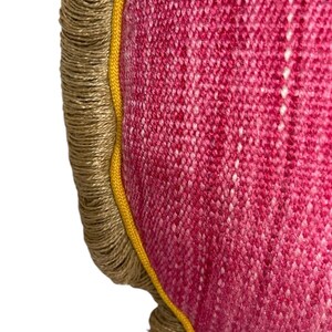 Puede incluir: Primer plano de una almohada redonda de color rosa con ribete amarillo y flecos marrones. La almohada est&aacute; hecha de una tela texturizada.