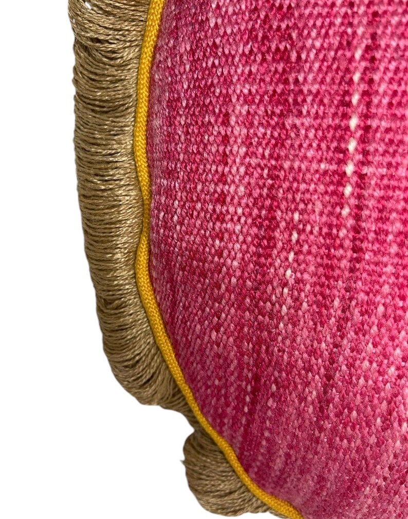 Puede incluir: Primer plano de una almohada redonda de color rosa con ribete amarillo y flecos marrones. La almohada est&aacute; hecha de una tela texturizada.