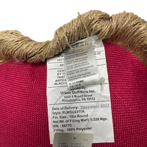 Puede incluir: Un coj&iacute;n redondo de color rosa con un ribete de tela marr&oacute;n. El coj&iacute;n tiene una etiqueta blanca adjunta con el texto "Urban Outfitters Inc. 5000 S Broad Street Philadelphia, PA 19112 Date of Delivery: 'December-2022 Style: PLWSU2311A Fin. Size: 18in Round Net Wt. of Filling Mat'l: 0.520 Kgs. RN.: 66170 Filling - 100% Polyester"