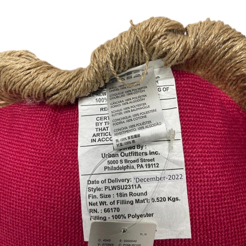 Puede incluir: Un coj&iacute;n redondo de color rosa con un ribete de tela marr&oacute;n. El coj&iacute;n tiene una etiqueta blanca adjunta con el texto "Urban Outfitters Inc. 5000 S Broad Street Philadelphia, PA 19112 Date of Delivery: 'December-2022 Style: PLWSU2311A Fin. Size: 18in Round Net Wt. of Filling Mat'l: 0.520 Kgs. RN.: 66170 Filling - 100% Polyester"