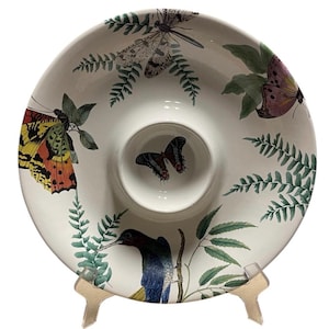 May include: White ceramic serving dish with a central dip bowl. The dish is decorated with colorful butterflies, dragonflies, and a hummingbird, along with green fern fronds. The dish is supported by a light-colored stand.