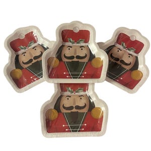 May include: A stack of five paper plates featuring a festive nutcracker design. Each plate has a red coat, black mustache, and a red hat with holly. The plates are white with a scalloped edge, perfect for holiday gatherings.