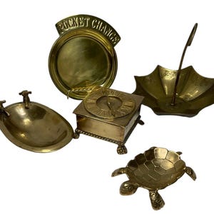 May include: A set of five brass trinket dishes. The dishes are shaped like a turtle, a clock, a small bathtub, an umbrella, and a round dish with the words "Pocket Change" on the top.