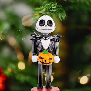 May include: A Jack Skellington nutcracker ornament featuring a white skull head with large black eyes, wearing a black and white pinstriped suit with a bat-shaped bowtie. The figure holds a small orange jack-o'-lantern and stands on a red base.