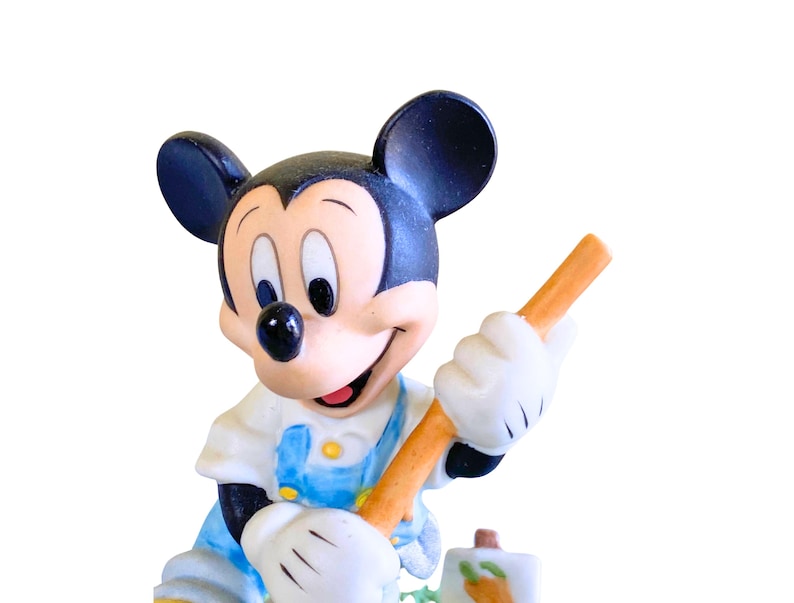 Peut inclure: Une figurine en c&eacute;ramique de Mickey Mouse portant une salopette bleue, tenant une carotte dans sa main droite. Il sourit et regarde vers la droite.