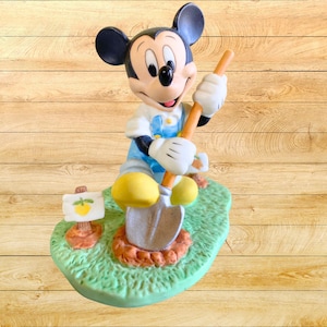 Peut inclure: Une figurine en c&eacute;ramique de Mickey Mouse habill&eacute; en jardinier, tenant une pelle et debout dans une zone herbeuse verte avec un petit citron jaune sur un panneau.