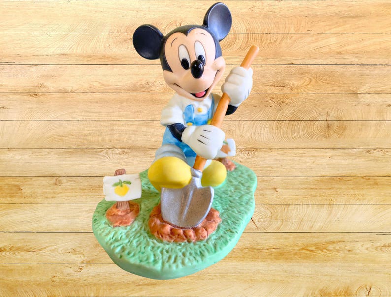 Peut inclure: Une figurine en c&eacute;ramique de Mickey Mouse habill&eacute; en jardinier, tenant une pelle et debout dans une zone herbeuse verte avec un petit citron jaune sur un panneau.
