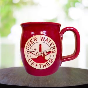 May include: Red ceramic mug with a white circle design featuring the text "Roger Waters" and "Us + Them". The circle also includes a graphic of two hands reaching towards each other.