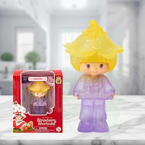 May include: A Strawberry Shortcake Almond Tea collectible figure with a translucent yellow flower-shaped hat and a purple glittery body. The figure holds a small tea set on a tray. It is packaged in a red box with the text Almond Tea.